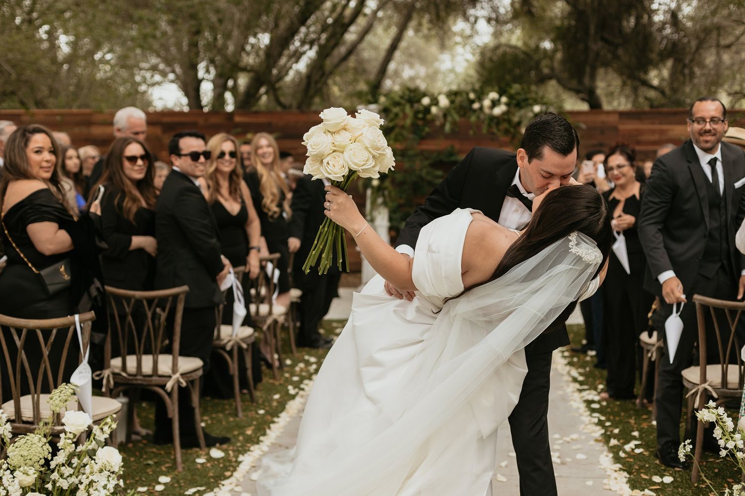 A romantic wedding ceremony moment with guests watching as couple shares a passionate kiss among scattered flower petals.