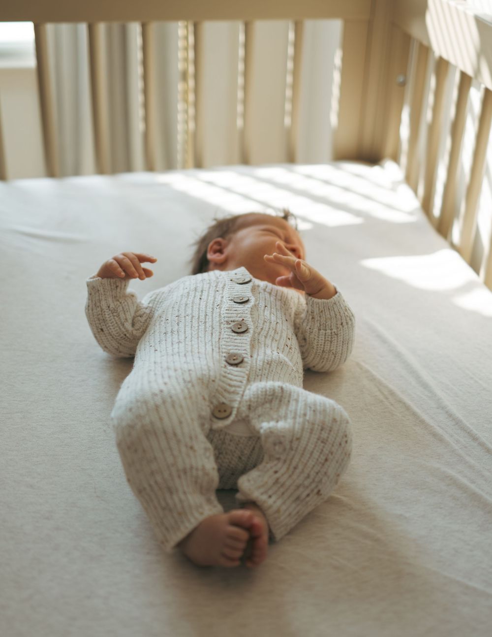 A sleeping newborn baby wearing a cozy knit onesie lies in a crib with sunlight streaming through the blinds.