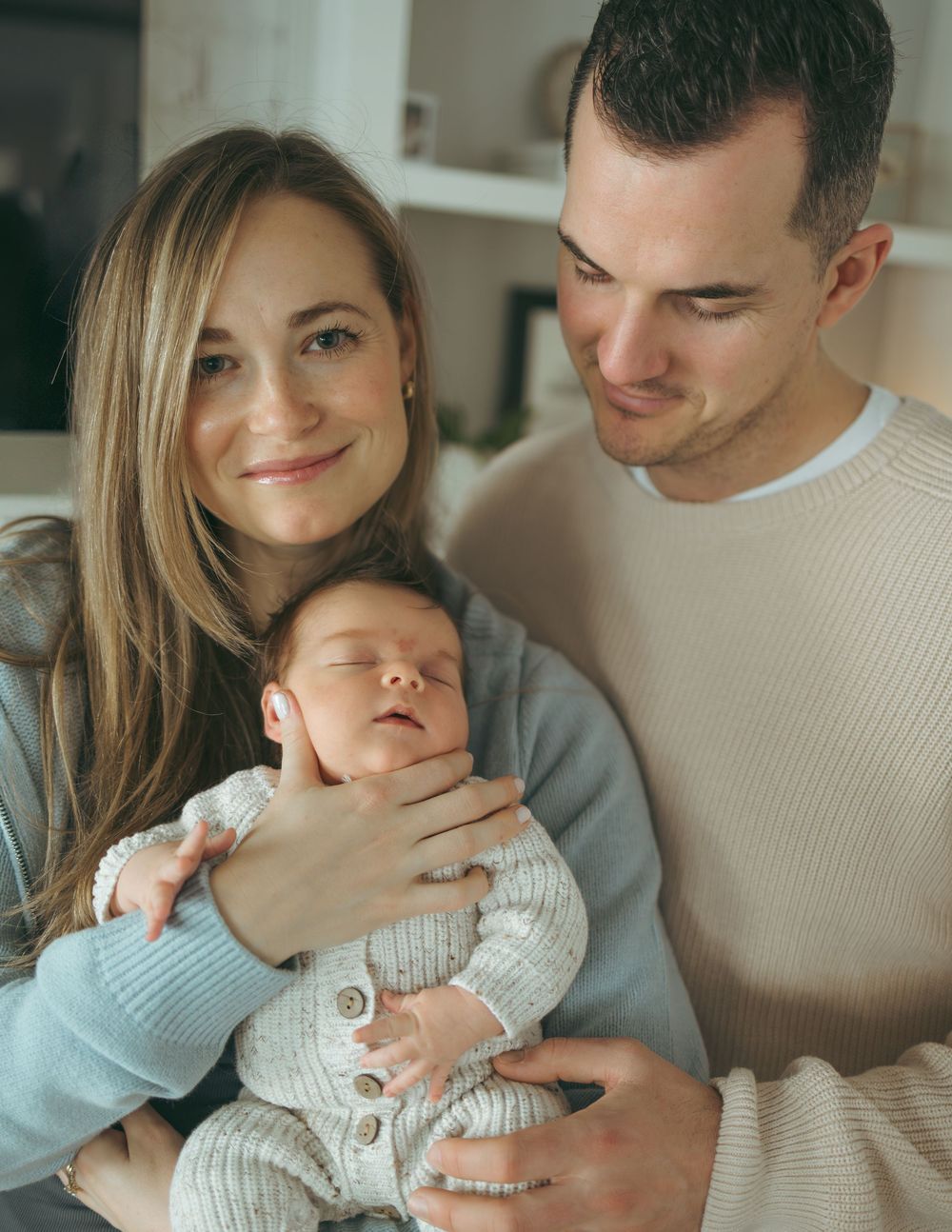 A couple in light colored sweaters share intimate moments while holding their baby in a bright indoor setting.