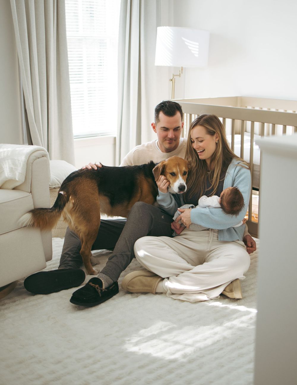 A couple relaxes on the floor with their beagle dog in a sunlit room with white curtains.