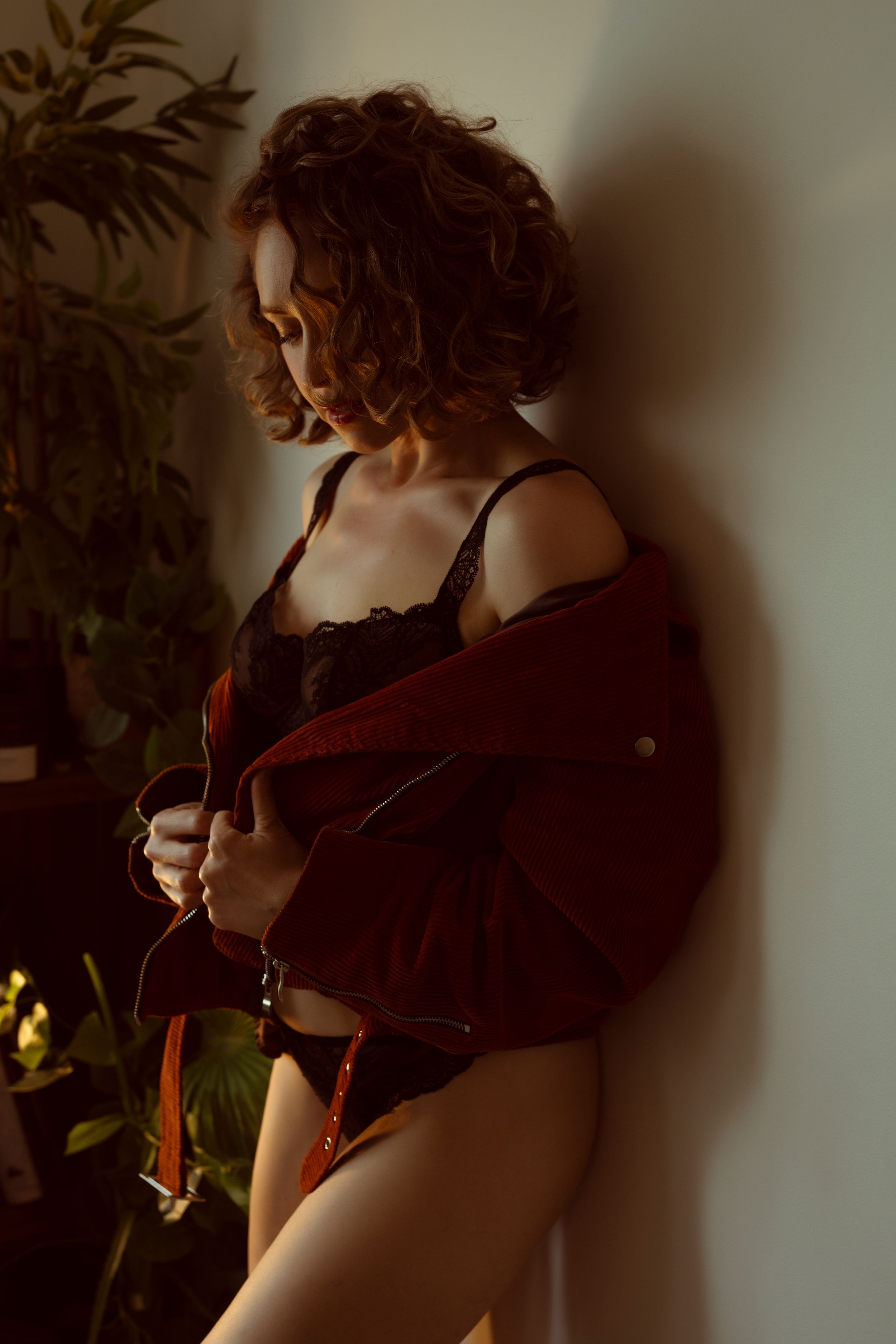 Person in red jacket and black lingerie poses against wall with dramatic shadows and indoor plant.