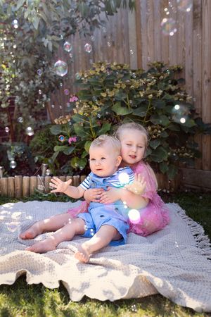 Two young children sitting on a white blanket outdoors while soap bubbles float around them in a garden setting.