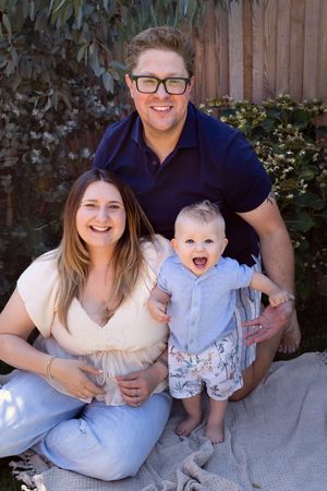 A happy family sits together on a grey blanket against a wooden fence in their backyard.