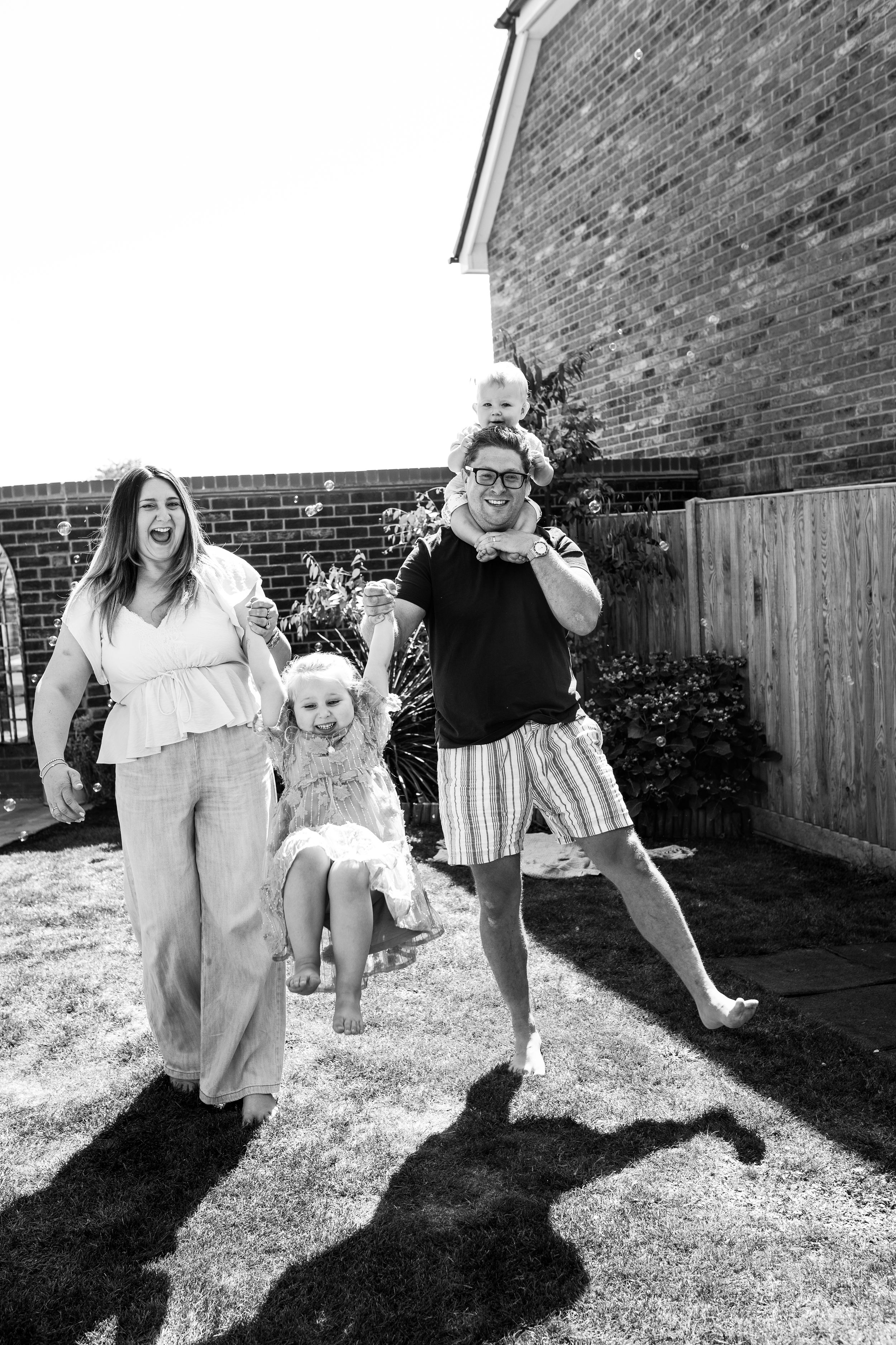 Family playing and jumping together in backyard creating shadows on brick wall and grass.