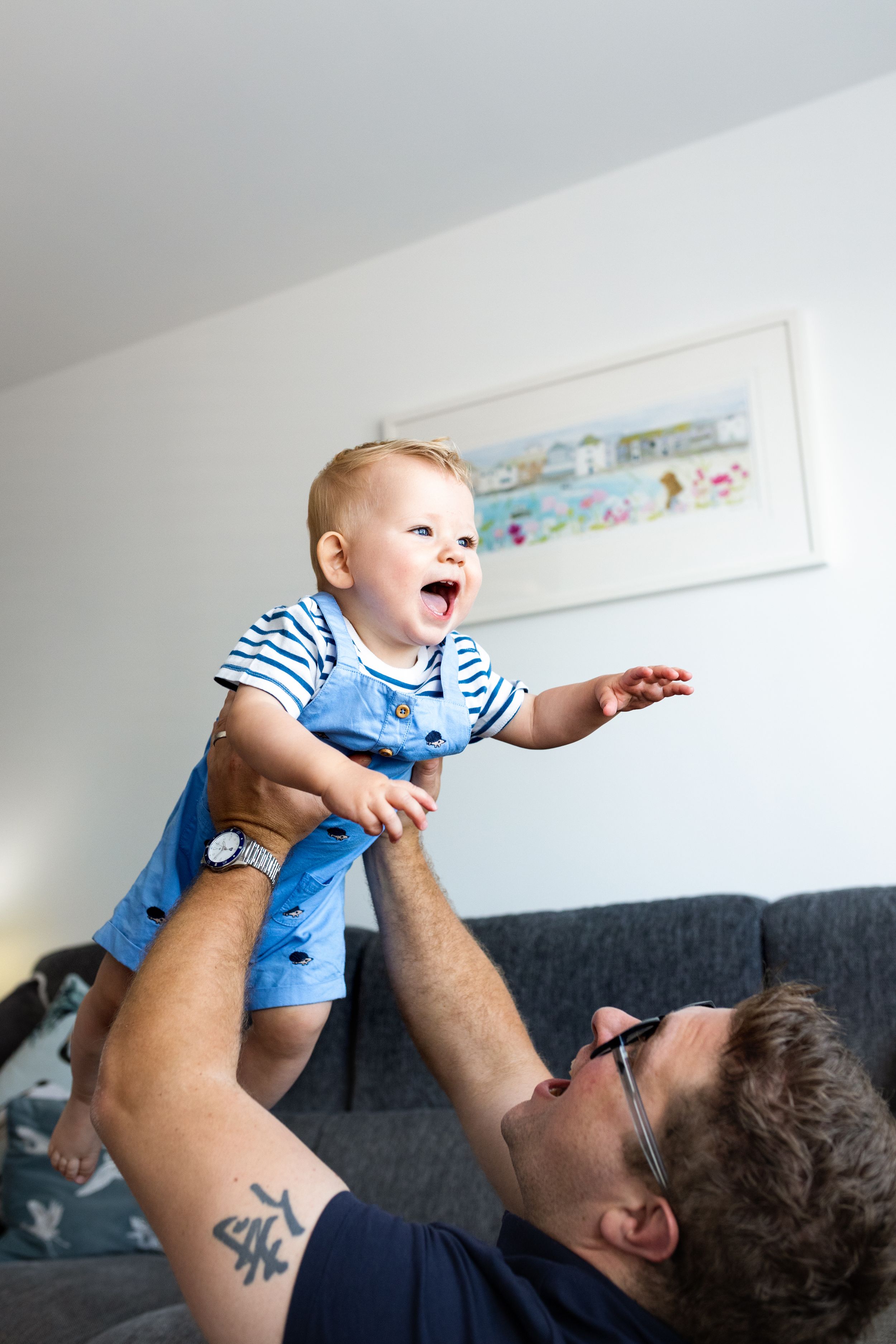 Baby in blue outfit being lifted playfully in the air against white walls.