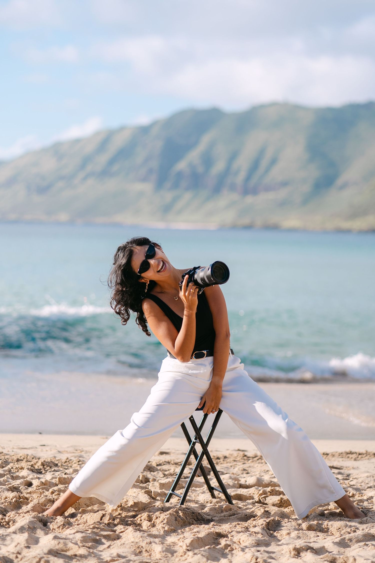Photographer in white pants sits on beach stool taking photos with mountains and ocean waves in background.