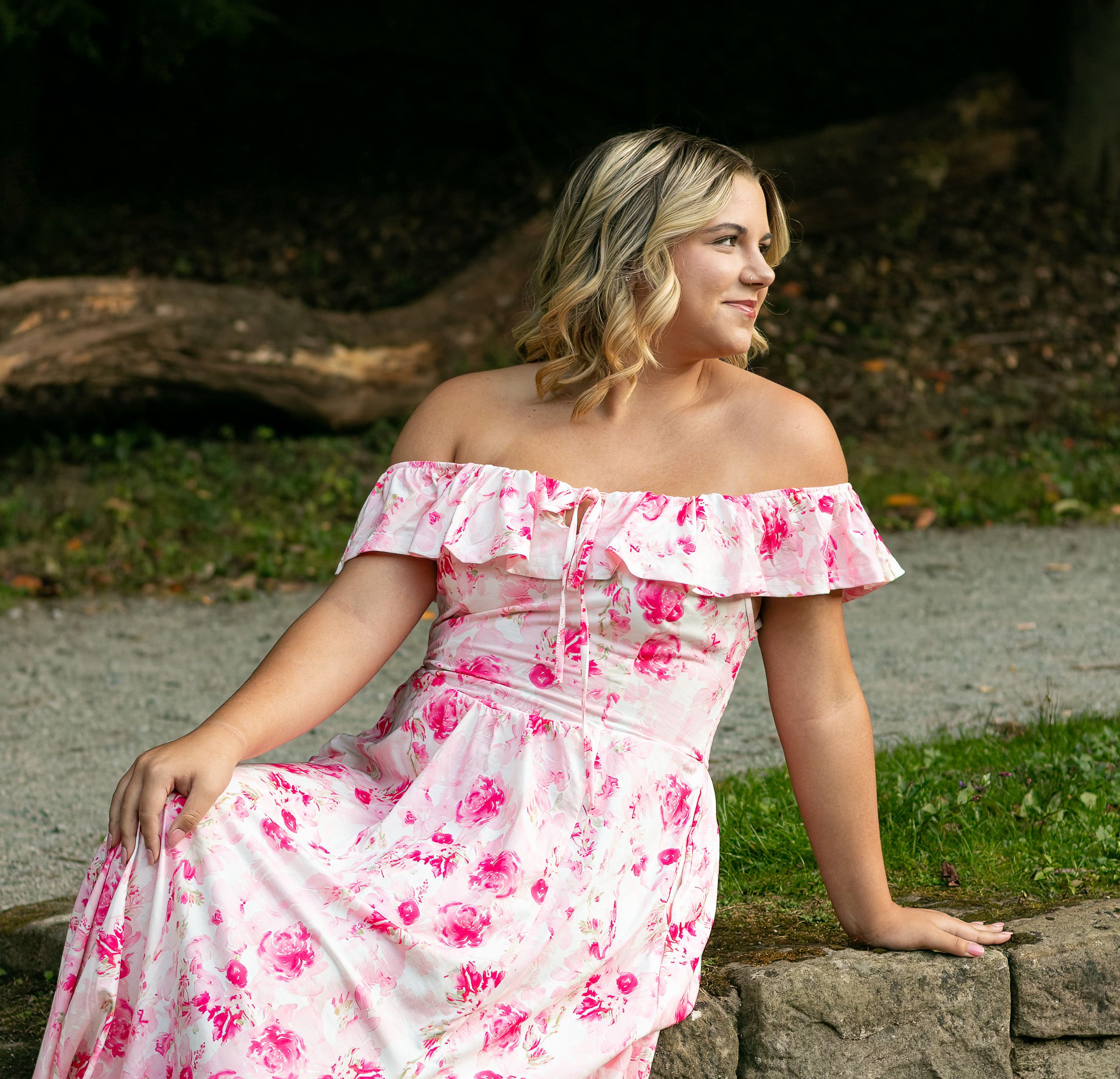 A woman in a pink and white floral off-shoulder dress poses outdoors against stone steps and greenery.