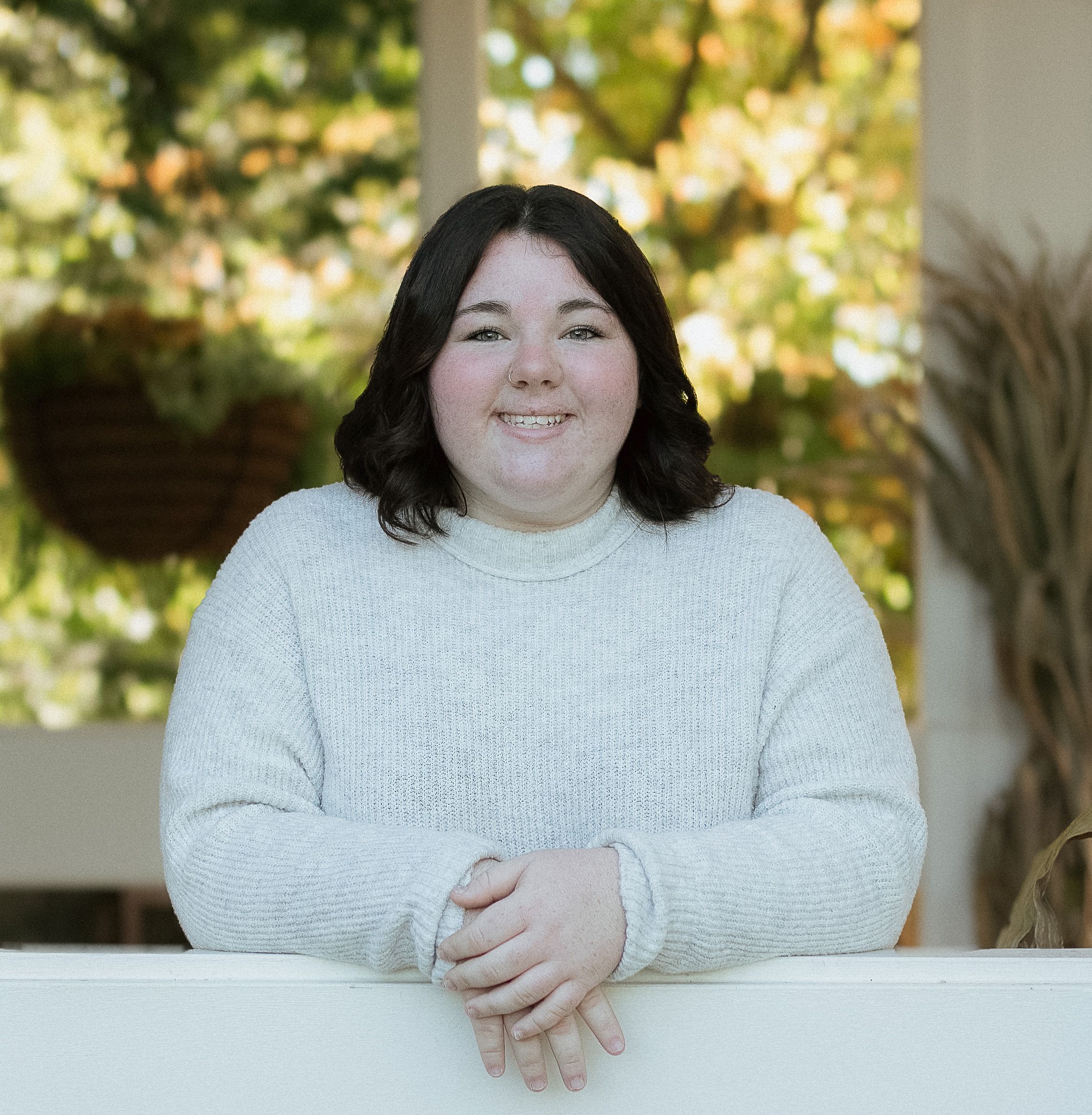 A person in a white sweater smiles warmly while sitting outdoors with autumn foliage in the background.
