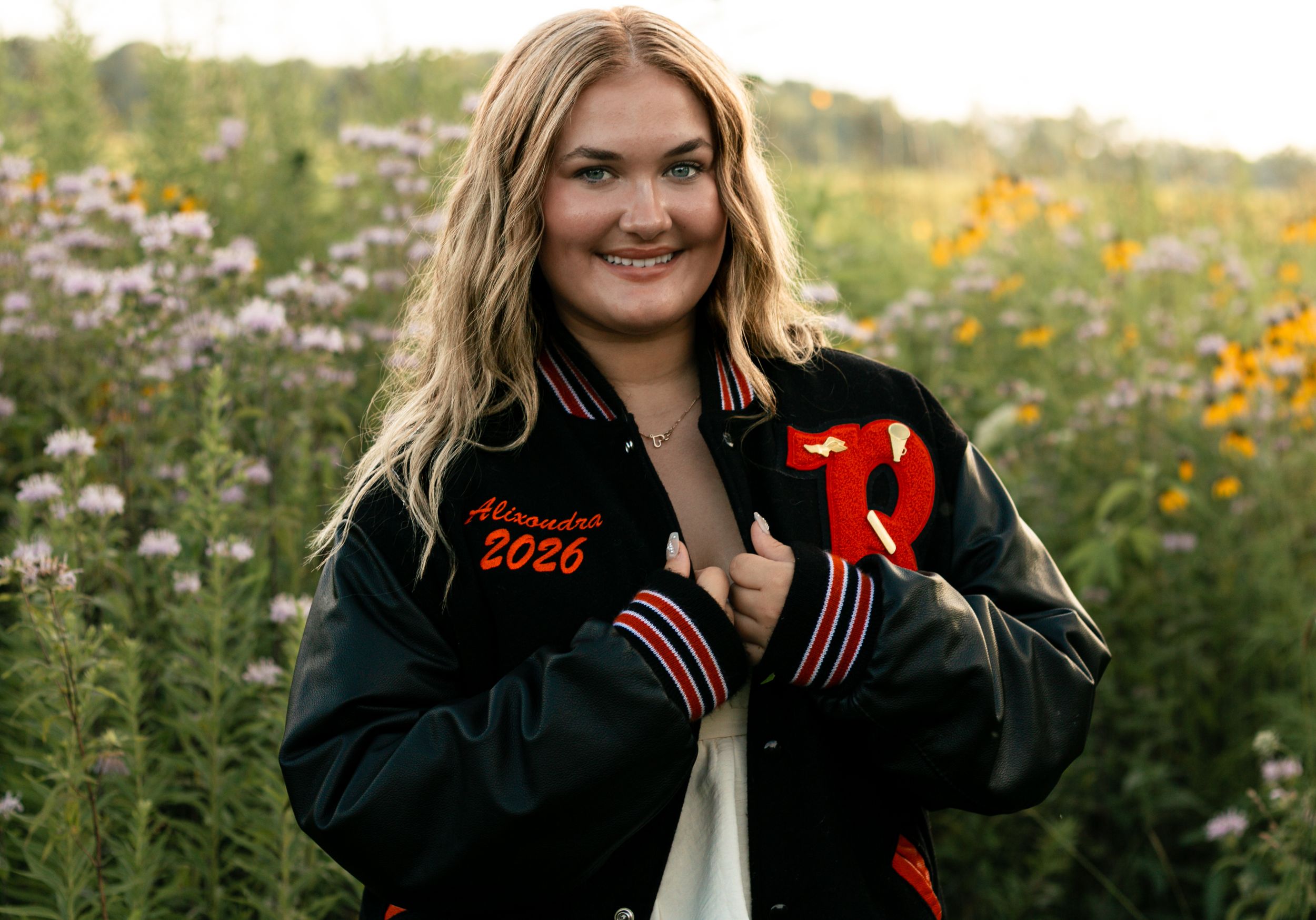 A person in a black and red varsity jacket poses in a field of wildflowers during golden hour.