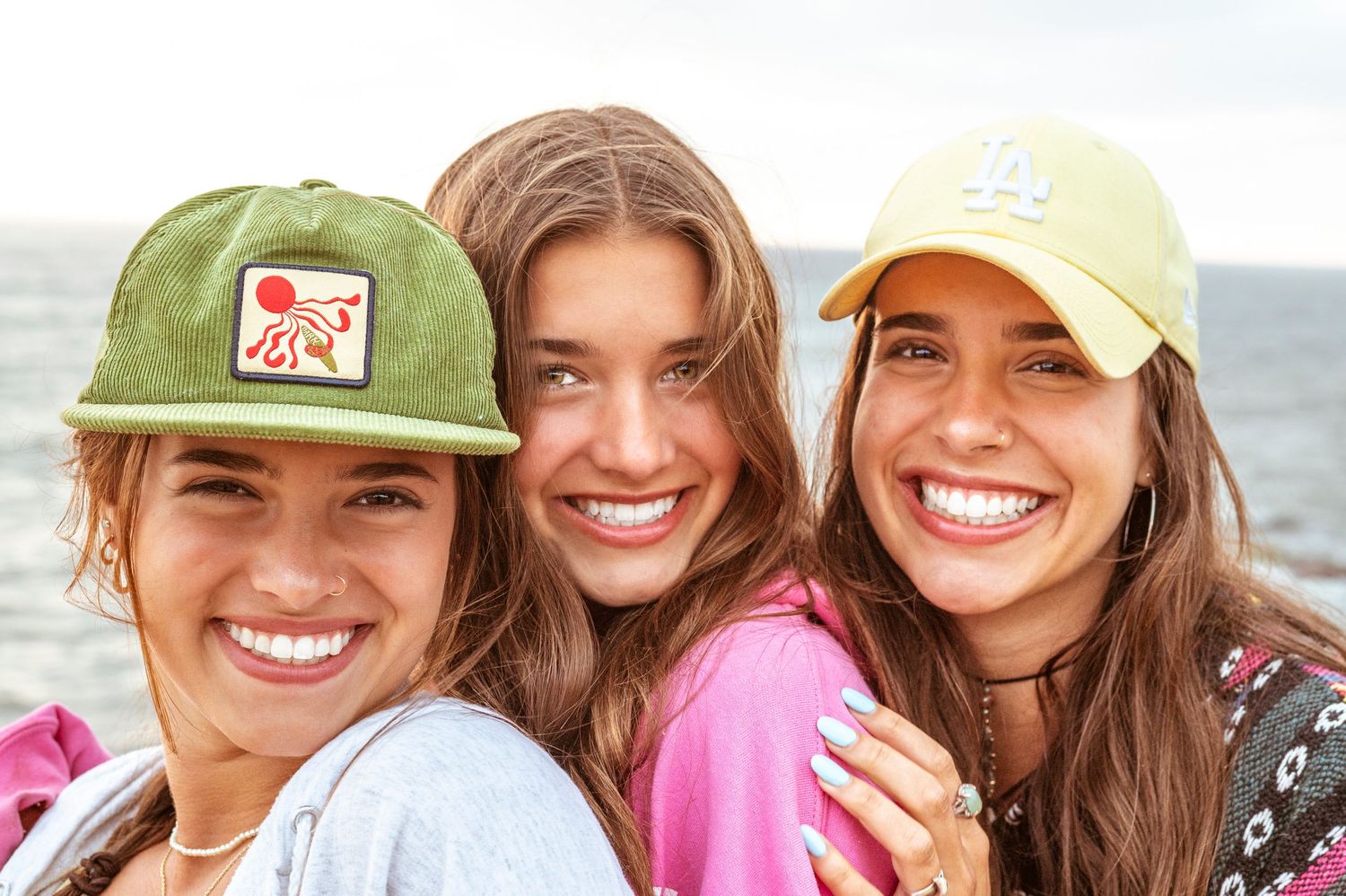 A group of friends wearing baseball caps smile together at the beach on a sunny day.