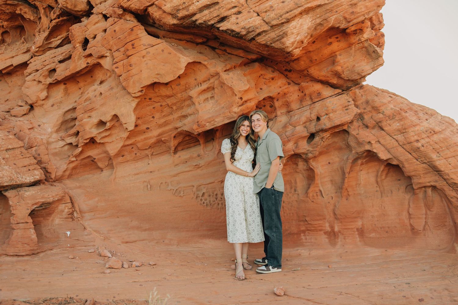 Desert engagement photoshoot with couple walking through sage brush against red rock formations in the distance.