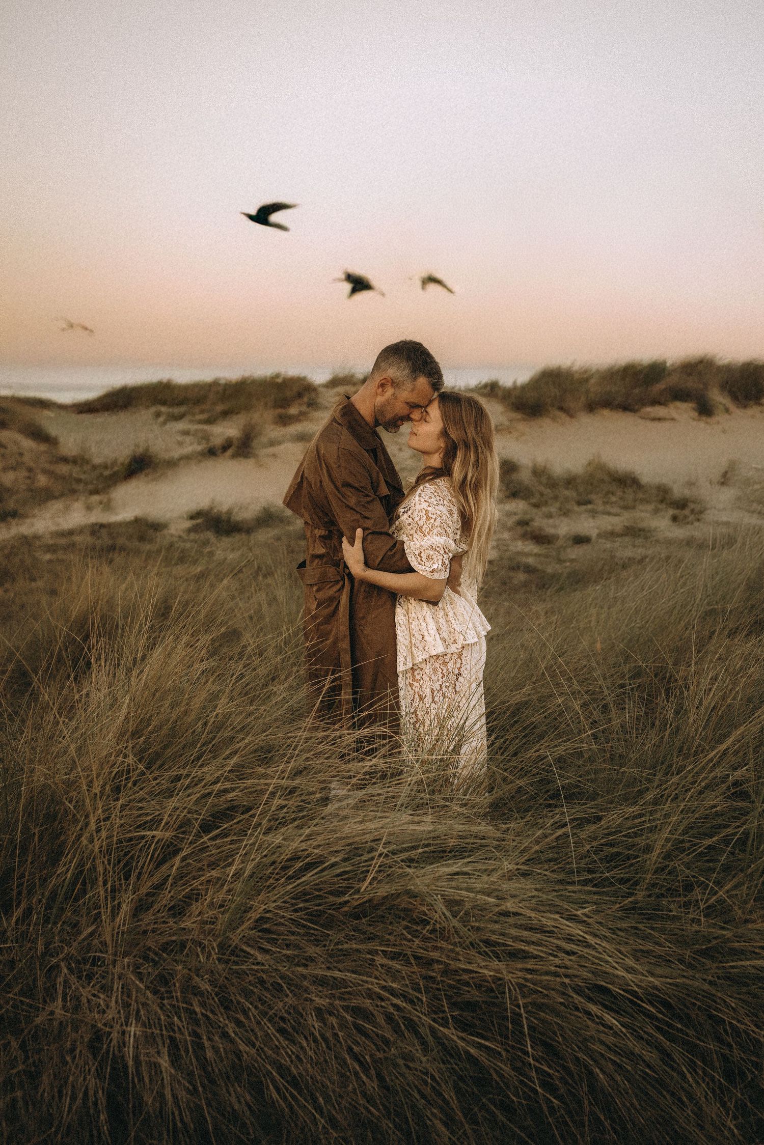 A romantic couple embraces in tall grass at sunset as birds fly overhead in the distance. Javea photographer