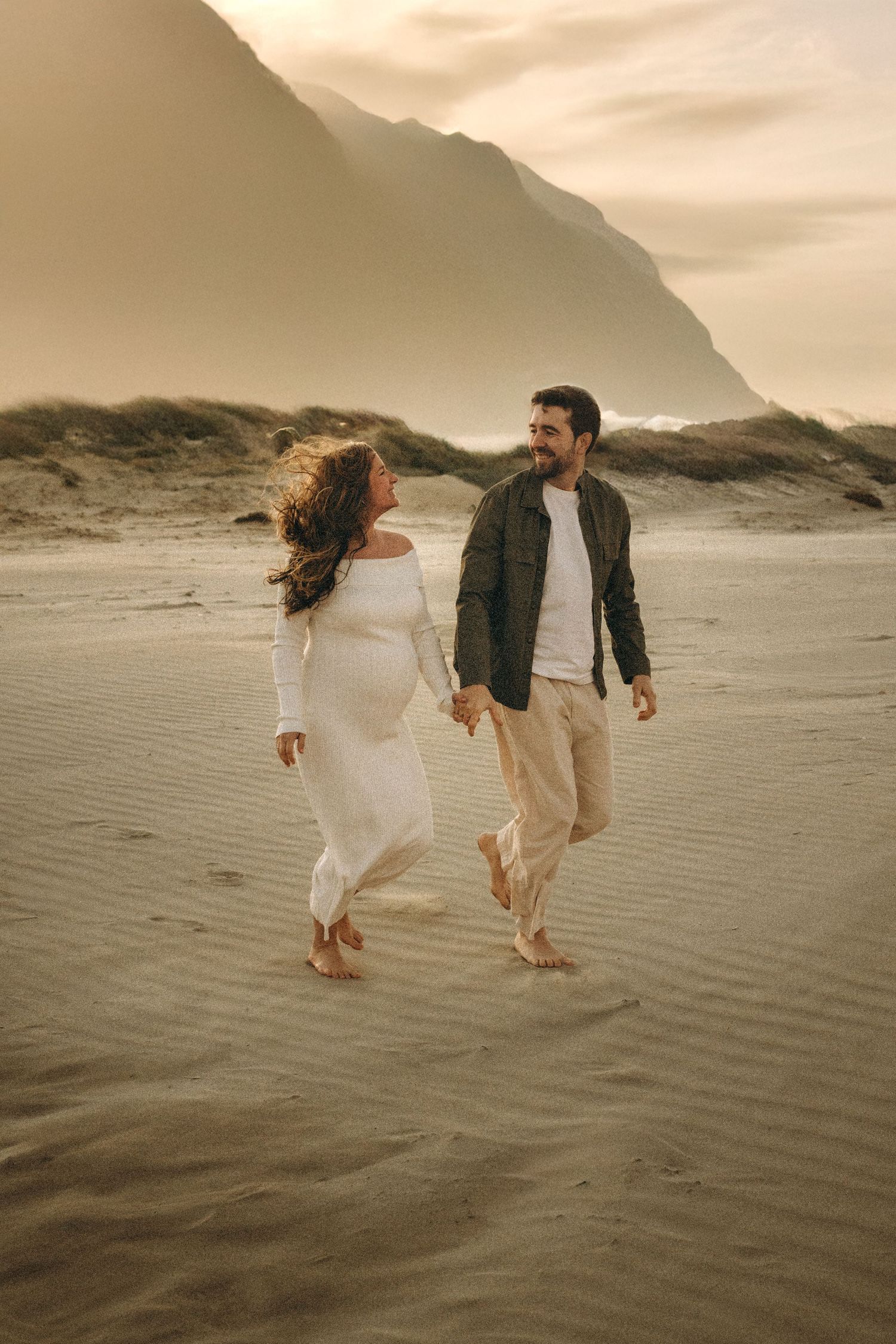 Two people walk barefoot on sandy beach with dramatic mountain cliffs and misty sunset backdrop in warm tones. Javea photographer