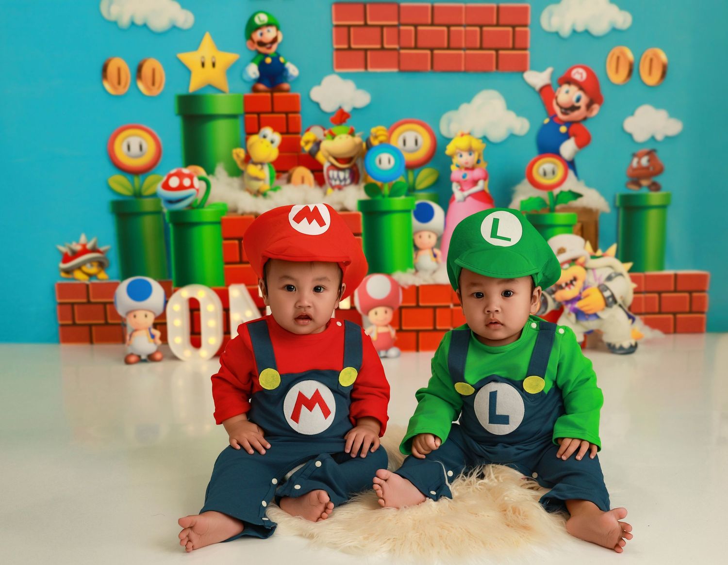 Two children wearing Mario and Luigi costumes pose in front of a Nintendo-themed backdrop with plush toys and pipes.