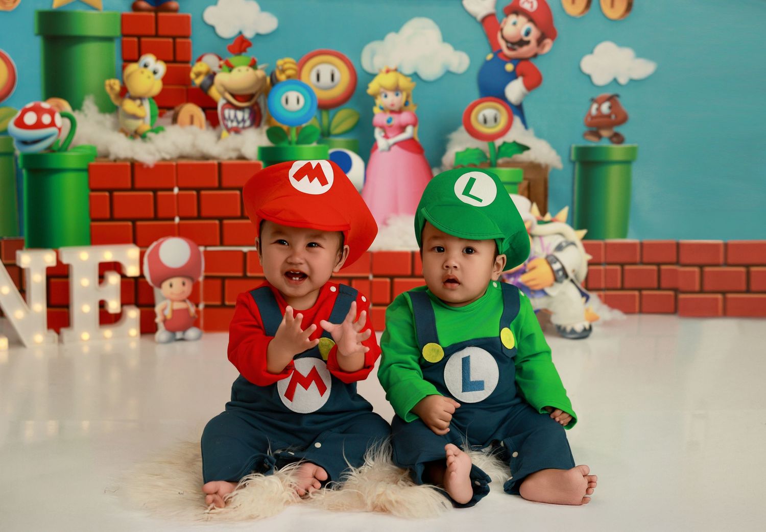 Two toddlers dressed as Mario and Luigi sit together against a Super Mario themed backdrop with pipes and blocks.