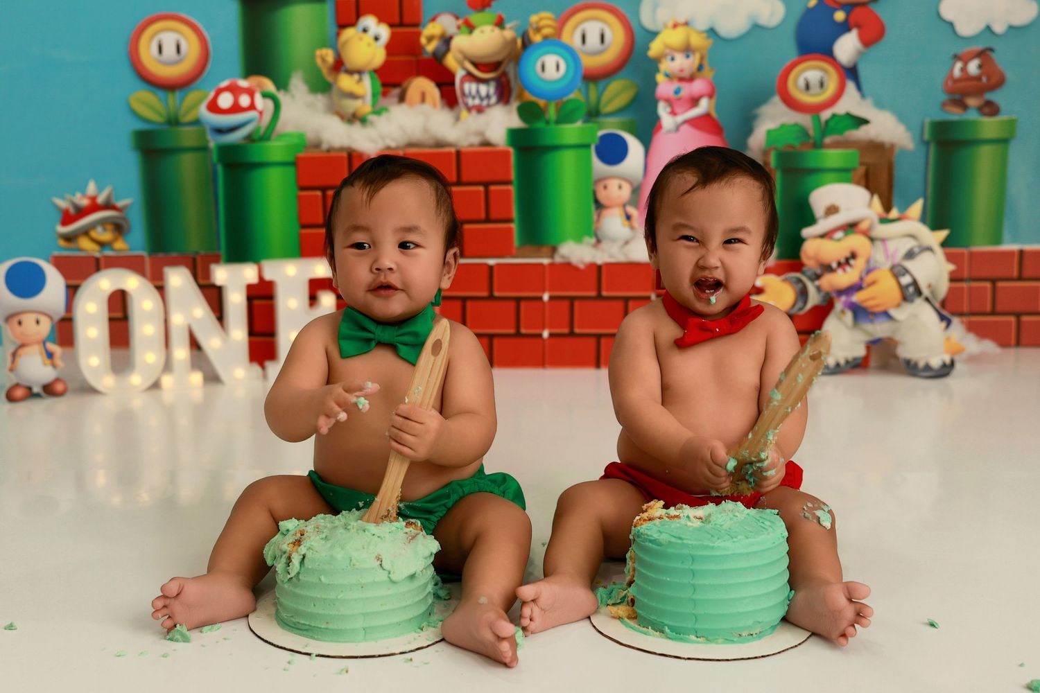 Twin babies posed against colorful Super Mario game backdrop with decorative cakes and gaming elements.