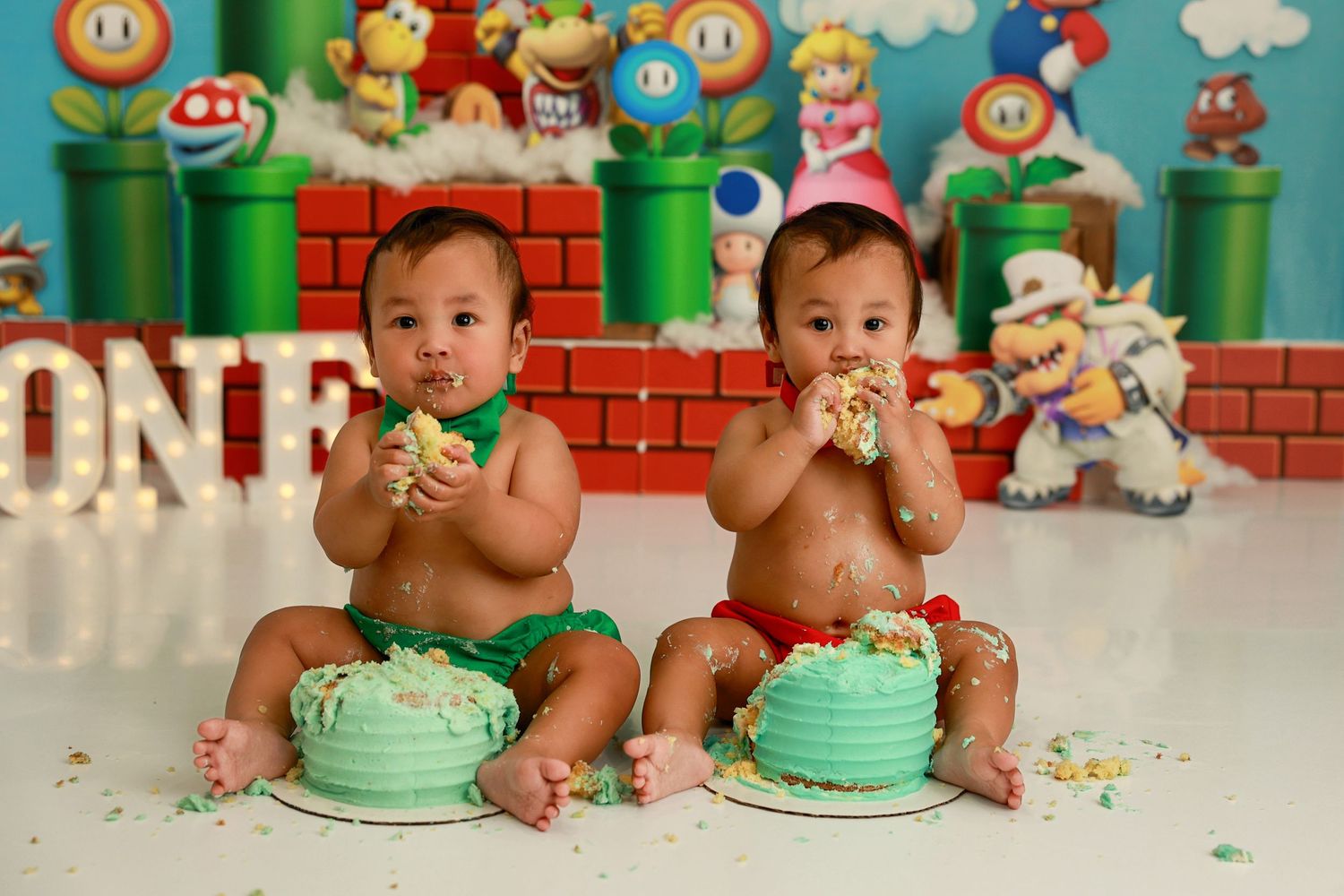 Twin babies in green bibs sit in front of matching smash cakes with Super Mario themed decorations in background.