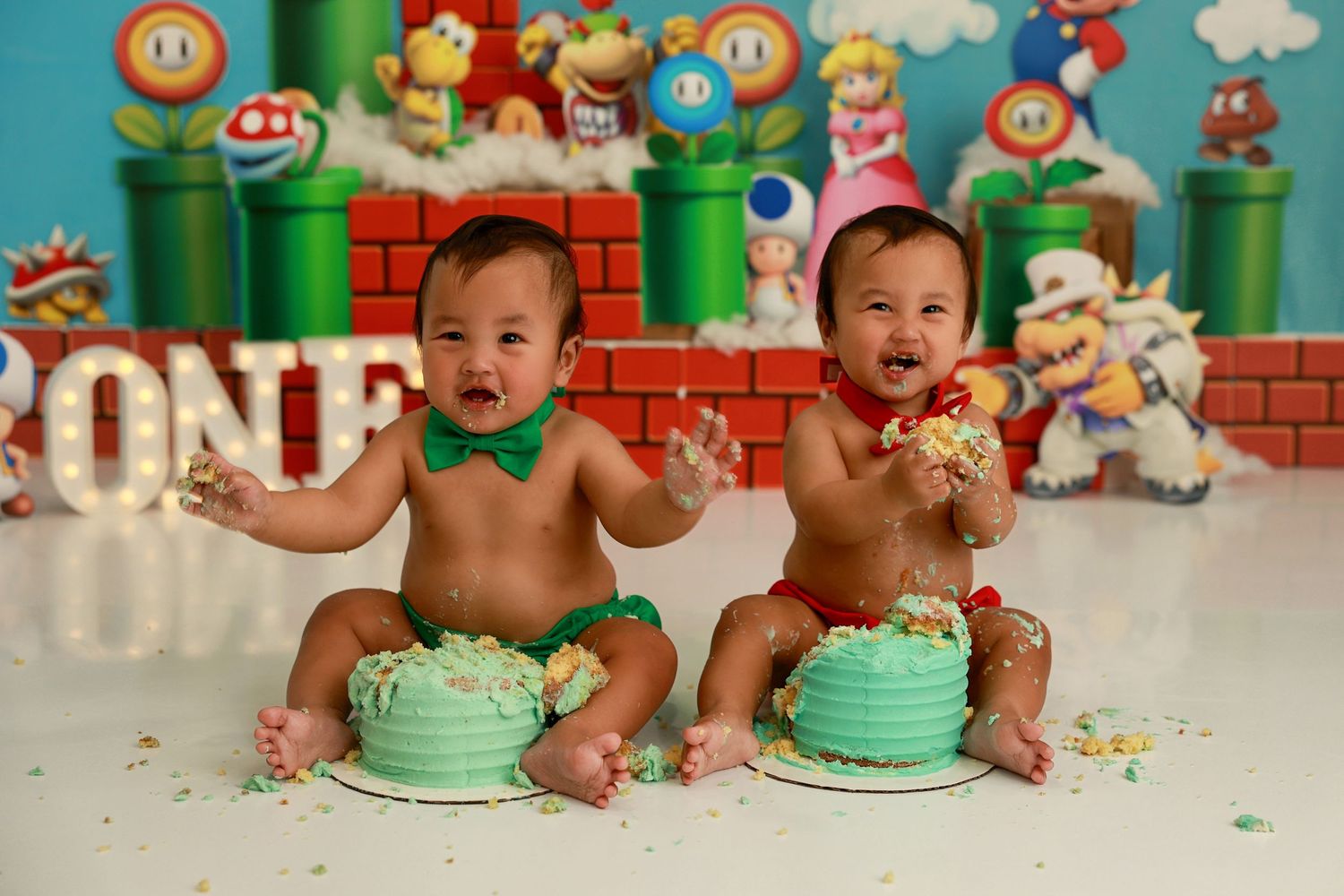 Smiling babies in Mario-themed outfits celebrate with smashed green cakes against gaming backdrop.