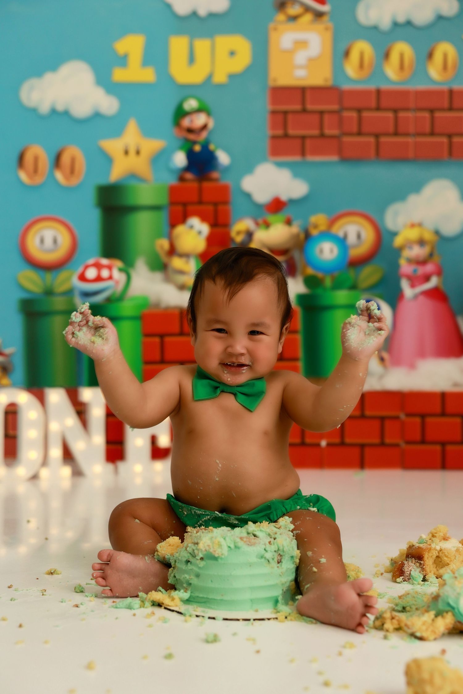 Baby wearing green outfit plays with smashed cake against Super Mario Bros themed background.