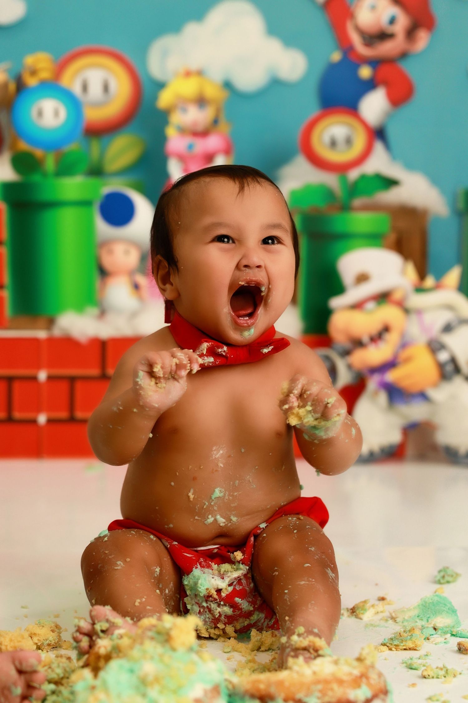 Joyful baby playing in cake and frosting with Super Mario themed backdrop featuring colorful game characters.