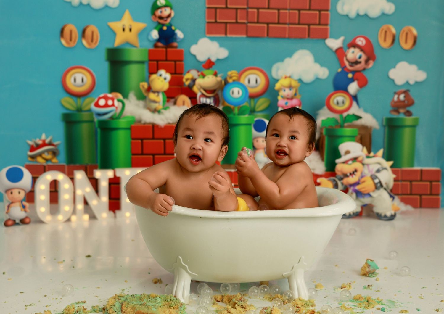 Two babies with matching green bibs prepare to eat their smash cakes against a Mario-themed backdrop.