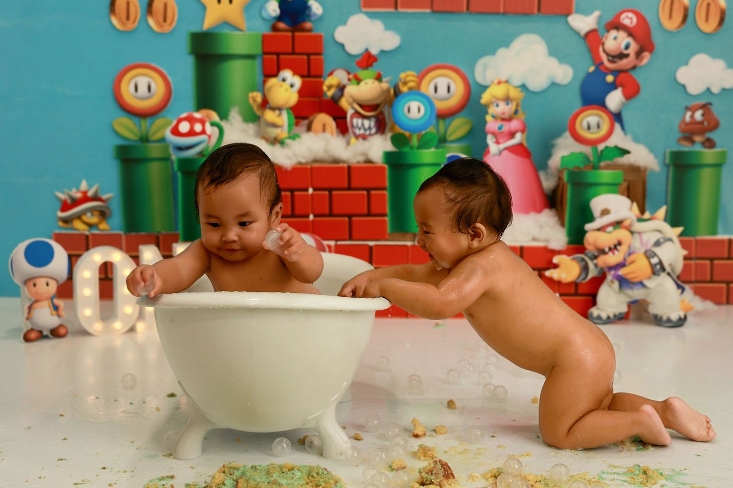 Two babies in mint green outfits enjoying cake smash photo session with Mario Brothers themed backdrop.