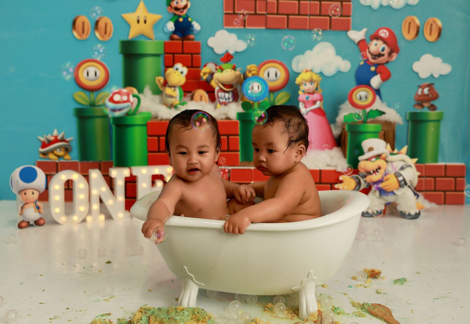 Baby sitting in white bathtub with Super Mario themed background and scattered cake pieces.