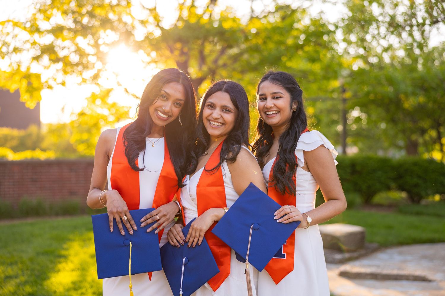 Three girls smiling with UIUC graduation attire at Hallene Gateway at sunset
