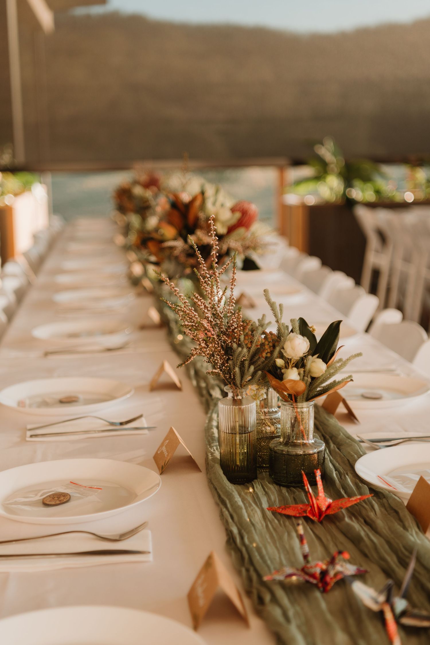 Elegant outdoor wedding reception table with floral centerpieces, origami cranes, and neutral place settings against a mountain backdrop.