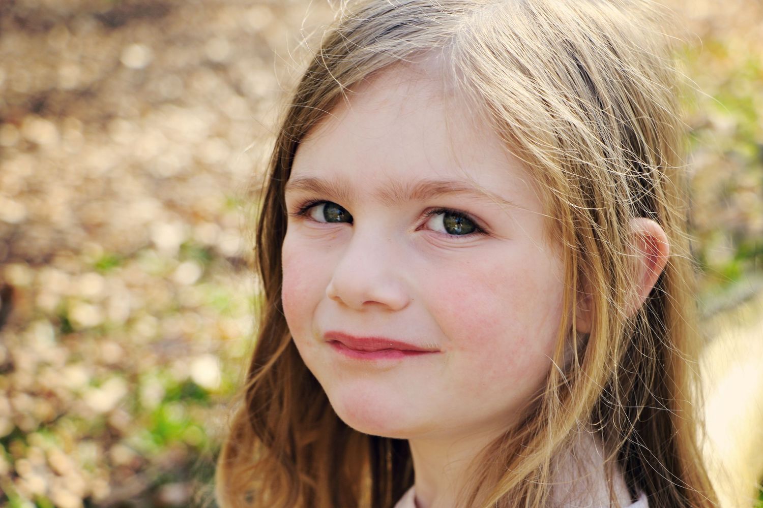 A young girl with long brown hair smiles softly while outdoors in natural lighting.