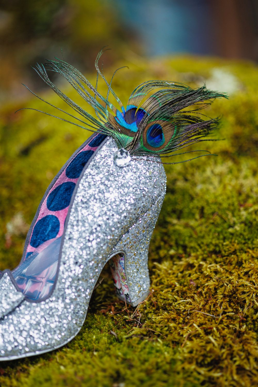 Glittery silver high heel shoe decorated with peacock feathers resting on mossy green surface.