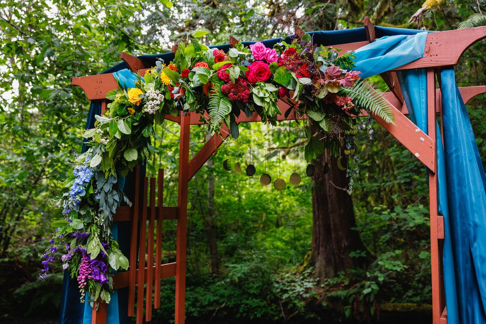 Colorful wooden wedding arch adorned with vibrant flowers and teal blue ribbon in forest setting.