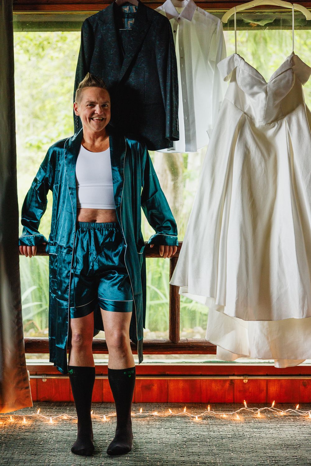 Person in teal satin robe standing before hanging wedding dress and tuxedo jacket before ceremony.
