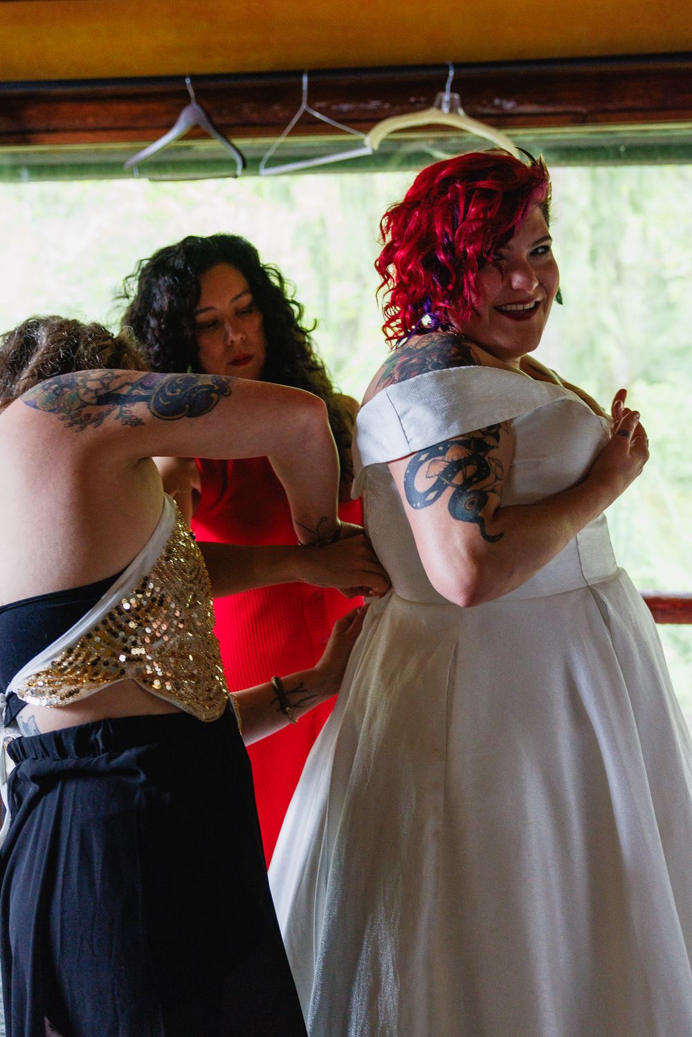 Bridesmaids helping red-haired tattooed bride into her white wedding dress before the ceremony.