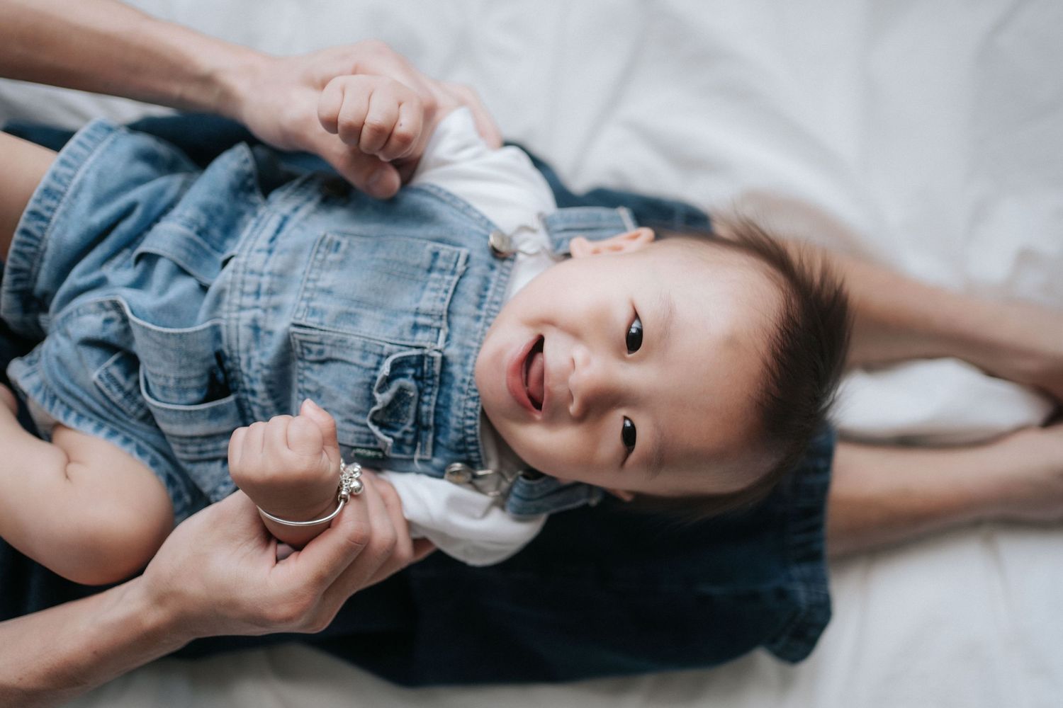 Smiling baby in denim overalls lying on white bed, held by adult hands in a joyful moment.穿著牛仔背帶褲的微笑嬰兒躺在白色床上，被大人抱著，享受著快樂的時光。