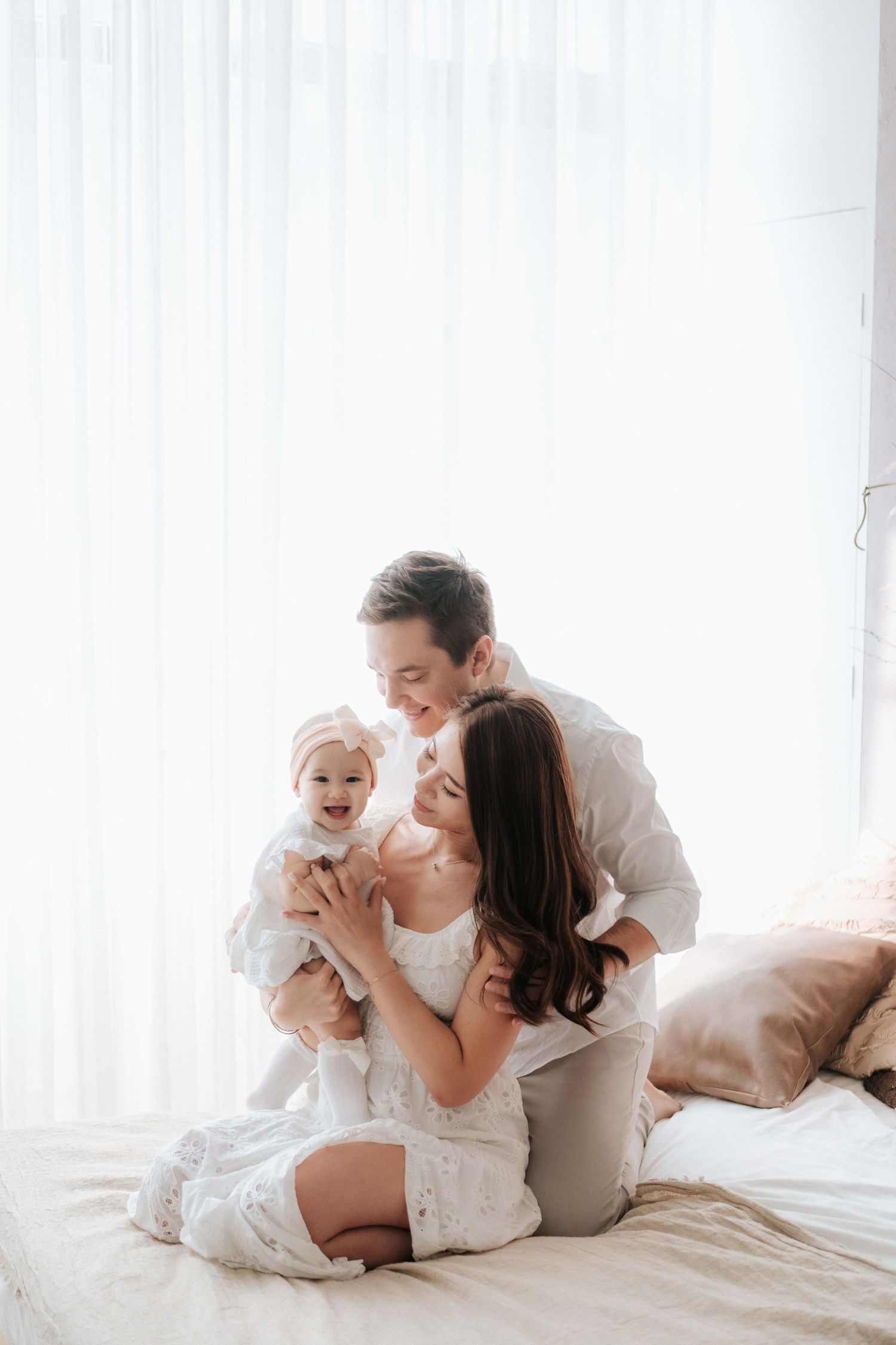 Happy young Asian family with mother, father, and baby in white outfits sharing a joyful moment on a bright bedroom bed.幸福的年輕亞洲家庭，媽媽、爸爸和寶寶穿著