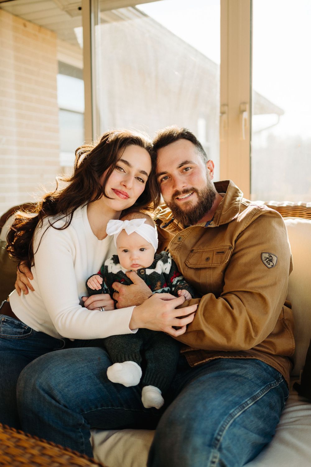 Family relaxing together on couch with baby in casual indoor setting filled with warm sunlight.