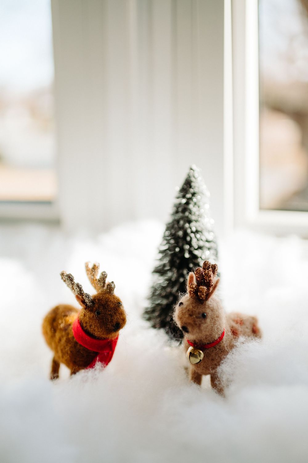 Two small felt reindeer decorations with red scarves next to miniature snow-covered tree.