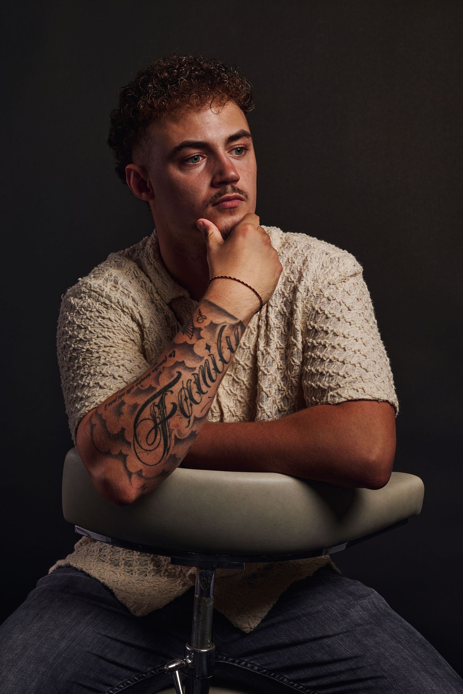 Jonge man met krullend haar en voorarm tattoo, zittend op een stoel in een nadenkende houding op een zwarte achtergrond.