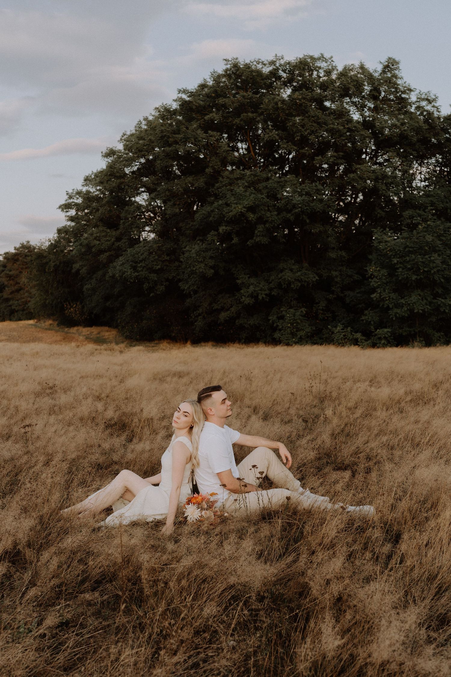 A couple sits in a golden field with their dog during sunset.