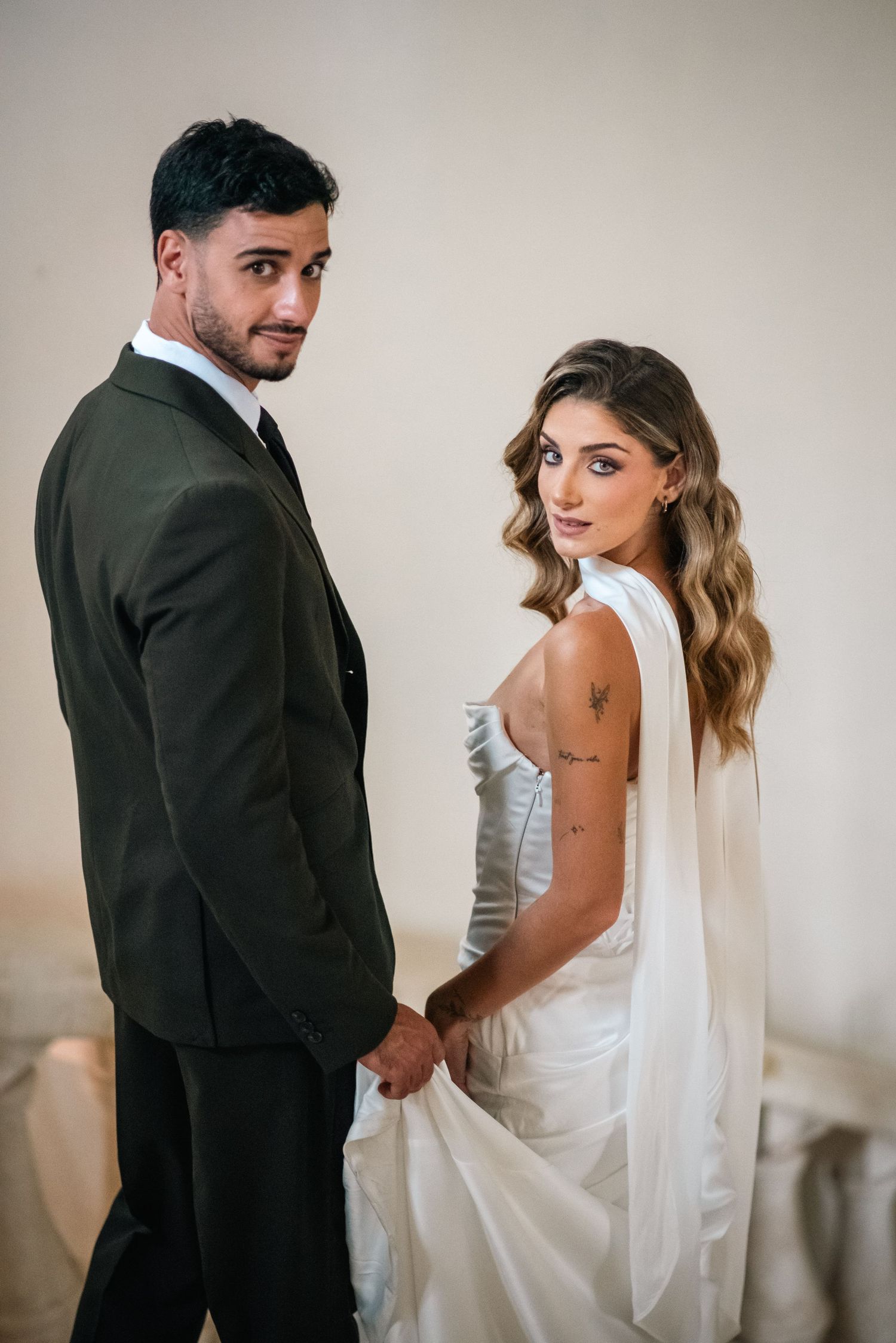 Elegant bride in white gown and groom in dark suit holding hands and posing for wedding portrait.