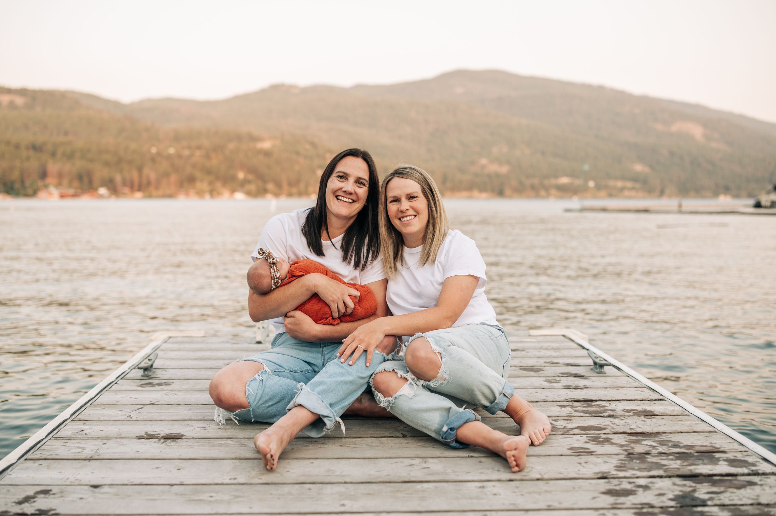 Two friends sitting cross-legged on wooden dock by lake holding small infant in orange blanket.