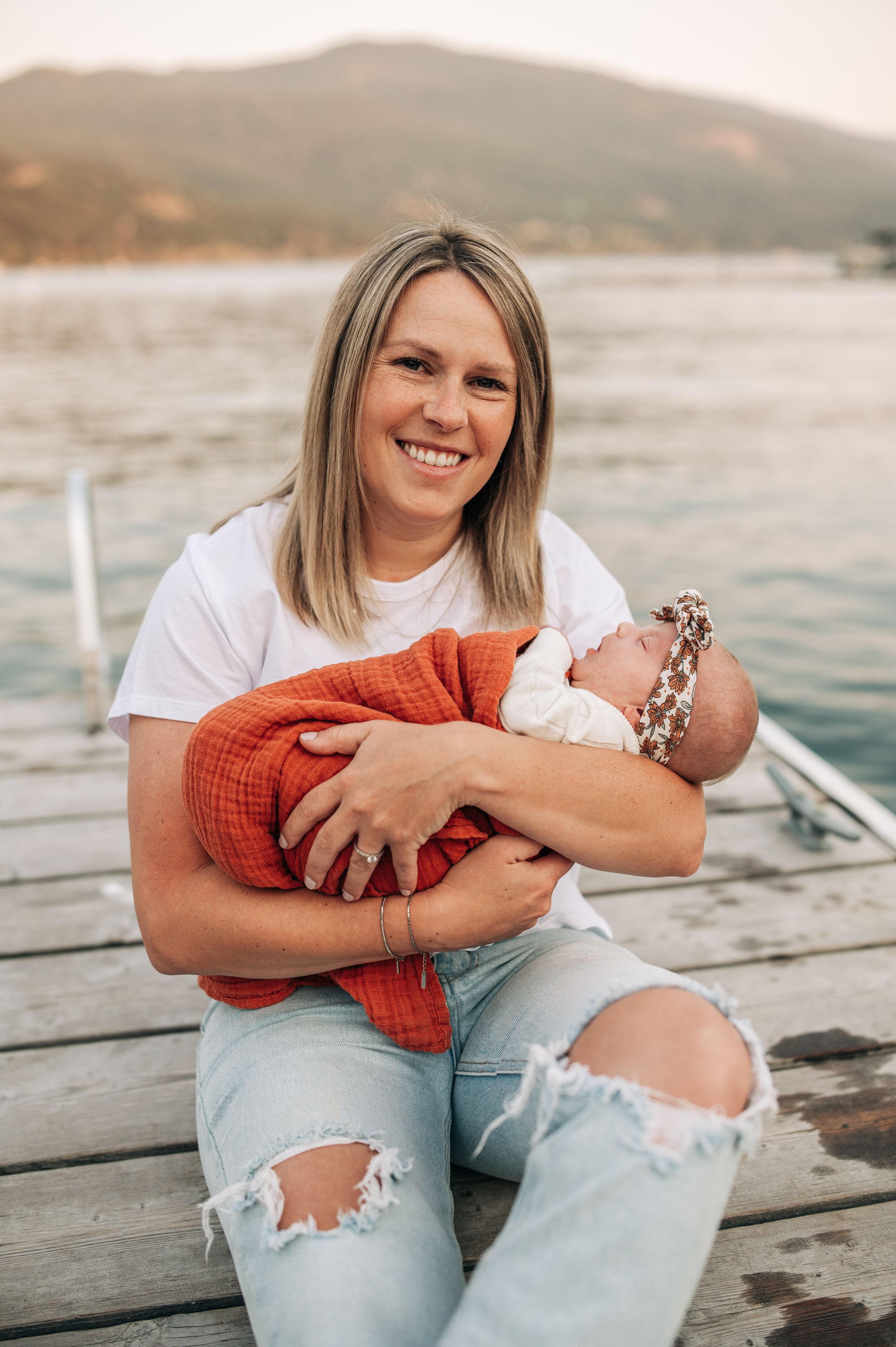 Person in ripped jeans holding baby wrapped in orange blanket on wooden dock by lake.