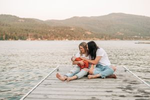 Two people sitting on dock with baby wrapped in orange blanket against mountain lake backdrop.