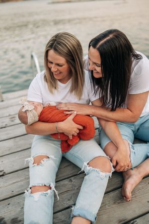 Two people in white shirts and ripped jeans sitting on dock with baby wrapped in orange blanket.