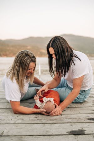 Two people in casual attire playing with infant wrapped in orange blanket on wooden dock.