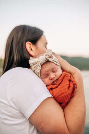 A sleeping newborn in an orange swaddle and bow headband being held against someone's shoulder.