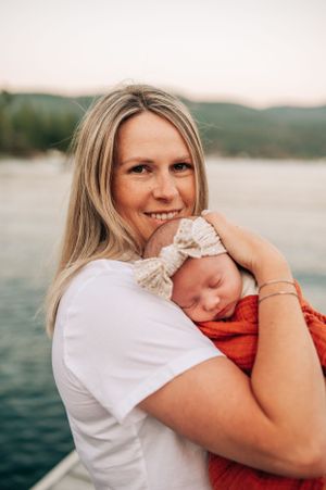 A joyful outdoor portrait with a sleeping newborn in orange wrap against a lakeside backdrop.