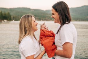 A candid moment of joy with a newborn in orange wrap against a mountain lake backdrop.