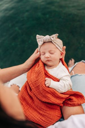 Sleeping baby wrapped in orange blanket wearing bow headband near water.